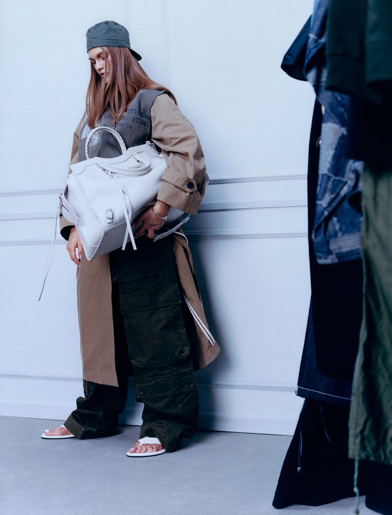 Woman in beige trench coat and green cap holding large handbag stands next to clothing rack indoors against a light blue wall.