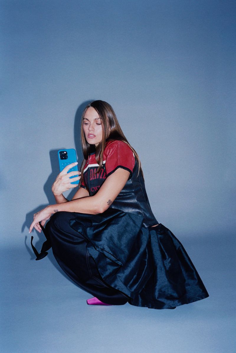Young woman in a sports jersey and skirt squats while holding a blue smartphone in a studio setting against a solid blue backdrop.
