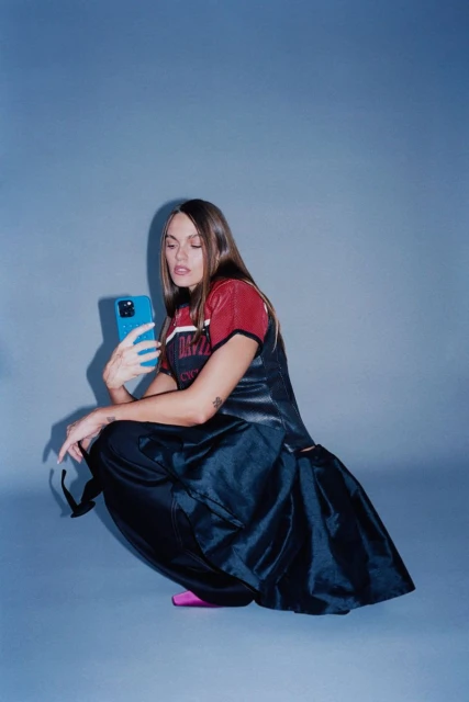 Young woman in a sports jersey and skirt squats while holding a blue smartphone in a studio setting against a solid blue backdrop.