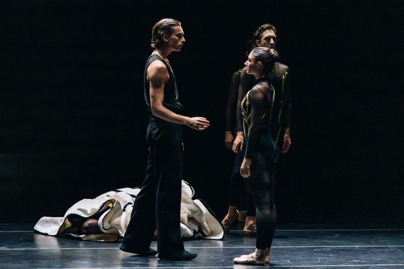 Three dancers in dark clothing on stage, one lying on the floor draped with a white and black patterned fabric. Low-key lighting.
