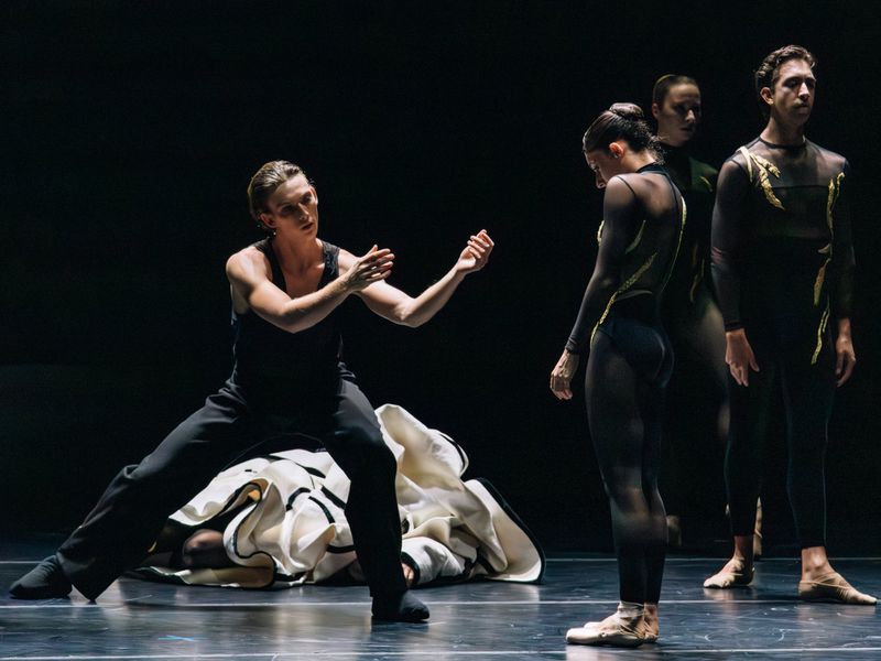 A group of ballet dancers on a dimly lit stage, one male dancer prominently positioned with arms raised in an expressive pose.