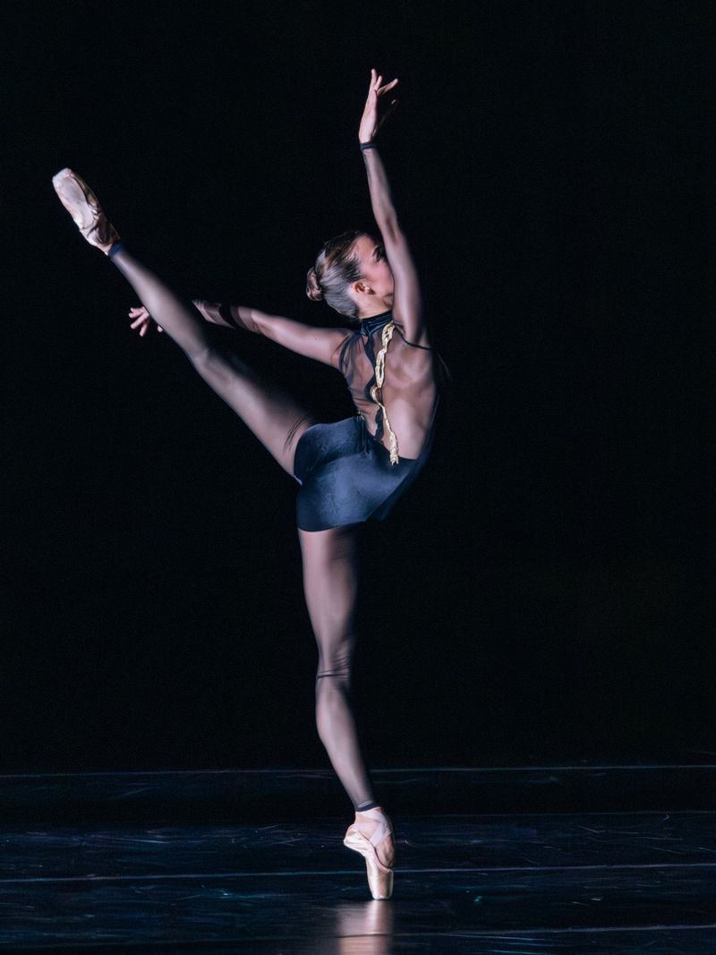 Ballet dancer in black leotard performs arabesque on pointe with dramatic lighting against a dark background, showcasing athleticism and artistry.