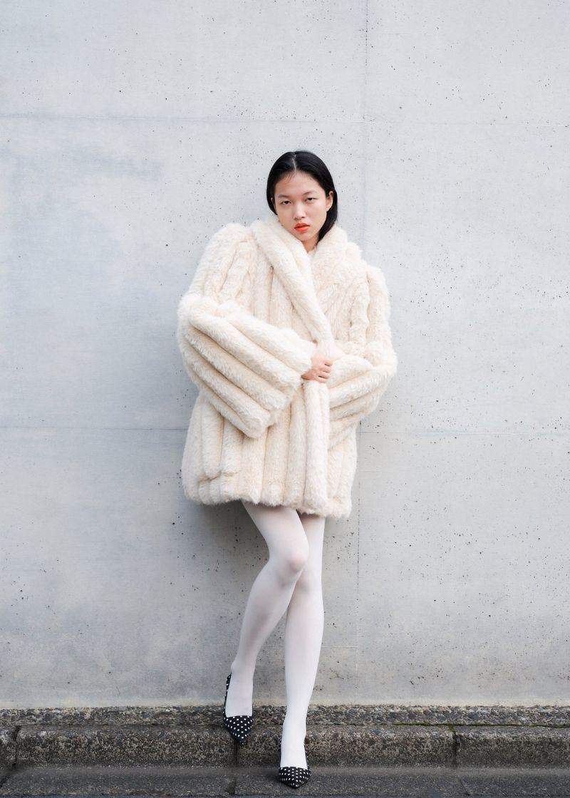 Asian woman in a fur coat, white tights, and polka-dot high heels poses against a textured grey wall in an urban setting.