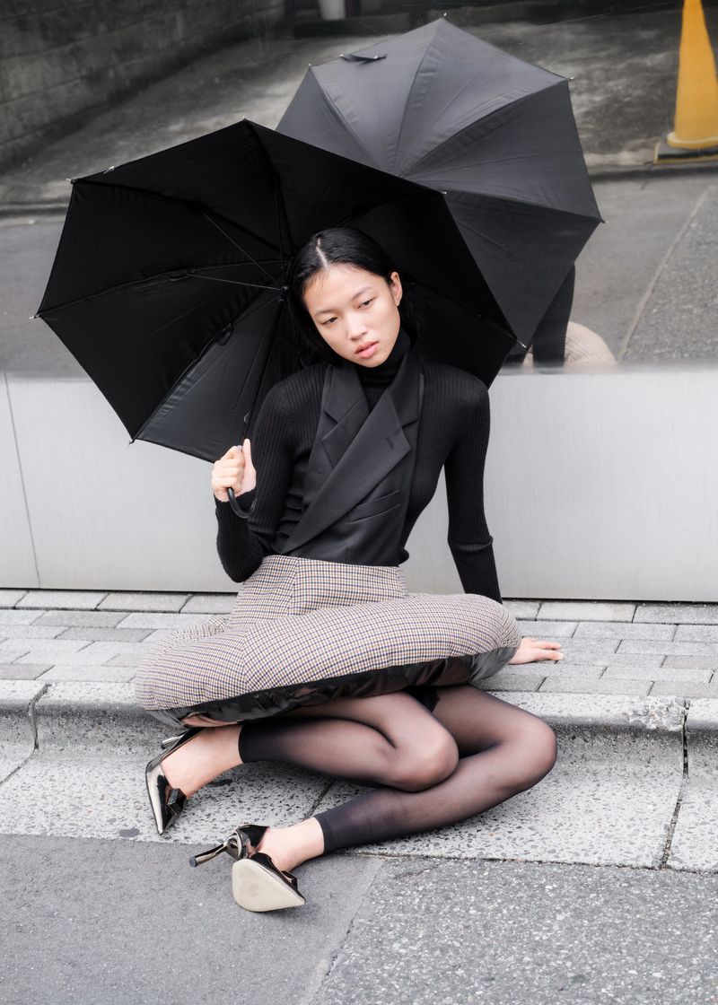 A woman in a skirt and top sits on stairs under a black umbrella on an overcast day in an urban outdoor setting.