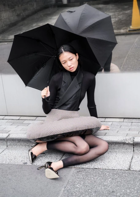 A woman in a skirt and top sits on stairs under a black umbrella on an overcast day in an urban outdoor setting.