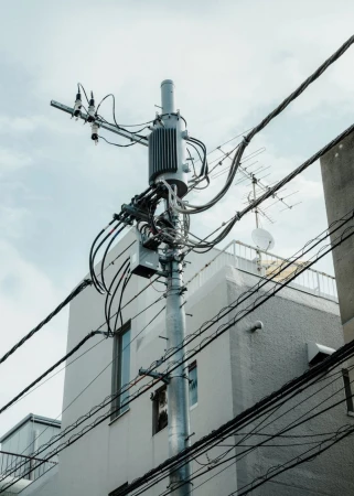 Utility pole with wires, cables, and hardware set against an urban backdrop with building exteriors and sky.