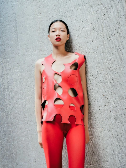 A fashionable Asian woman poses against a textured concrete wall in a cutout red top and matching red pants in a fashion editorial.