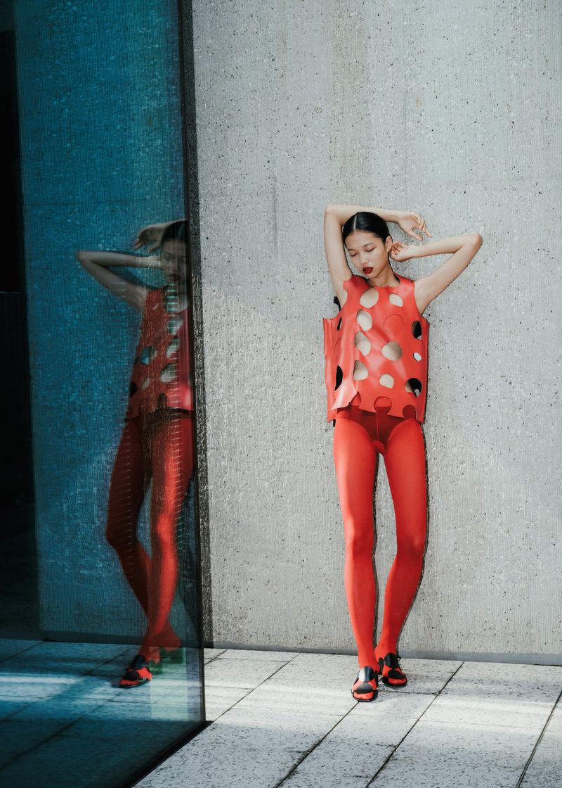 A woman in a red outfit poses against a textured wall and a glass panel reflecting her image, with sunlight enhancing the details.
