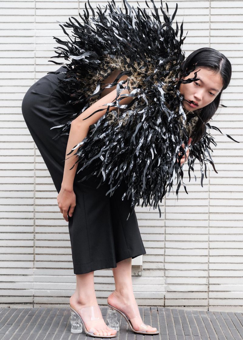 Adult female fashion model poses in a black outfit with a large feathered accessory against a tiled wall, wearing clear block heels.