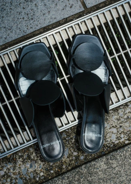 Top-down studio shot of black Pierre Hardy slide sandals with circular appliques, placed on a gray stone surface with a metal grate.