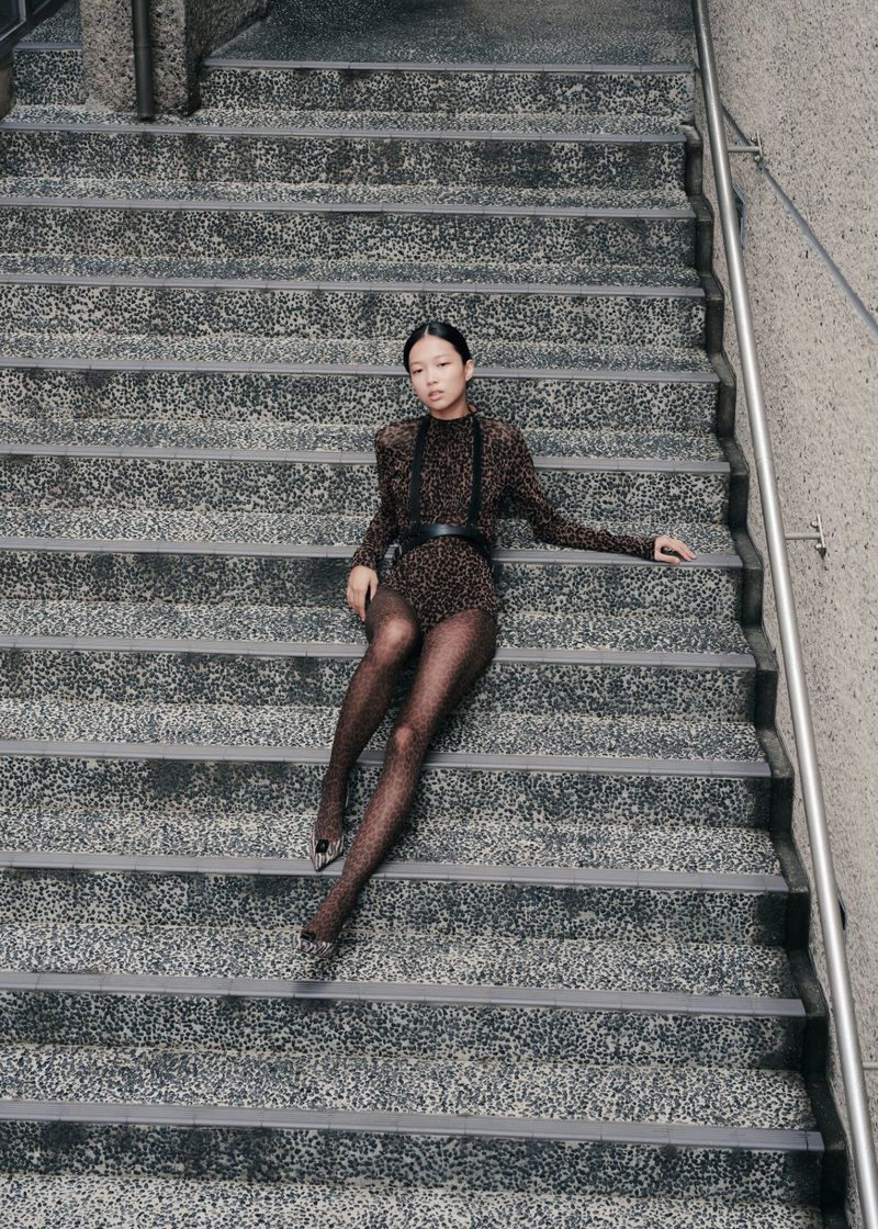Woman in leopard print bodysuit and sheer tights poses sitting on gray concrete steps in an outdoor urban setting, overhead shot.