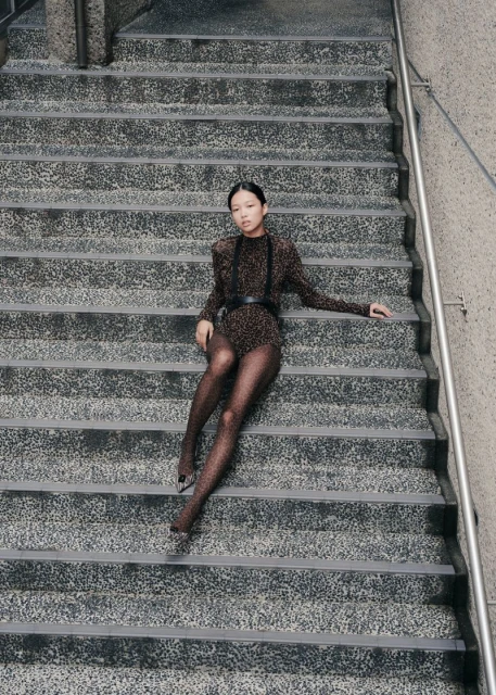 Woman in leopard print bodysuit and sheer tights poses sitting on gray concrete steps in an outdoor urban setting, overhead shot.