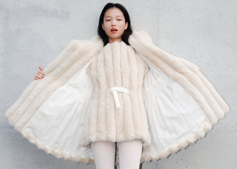Woman poses in an open, light-beige faux fur coat against a concrete background. The coat has a white lining and a tied waist.