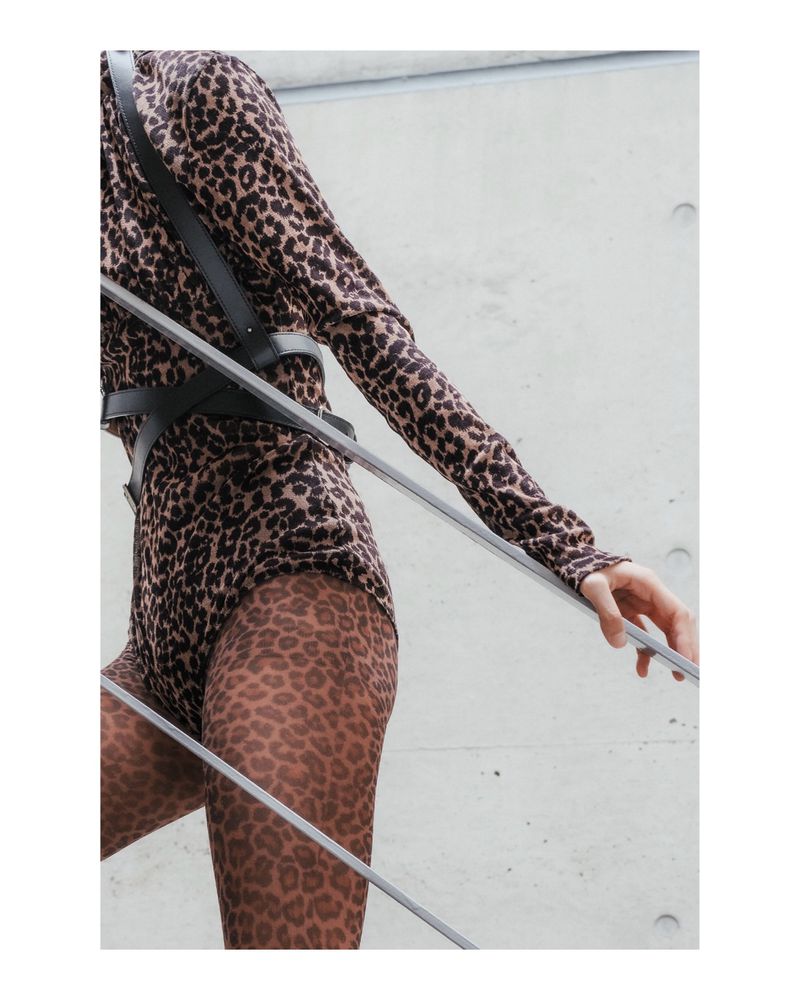 Fashion shot featuring a woman in a leopard-print bodysuit and tights leaning against metal bars in front of a concrete wall.