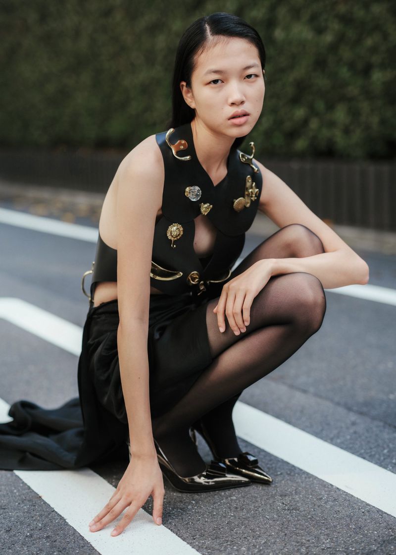 Young Asian woman poses crouching in a black fashion ensemble on a city street, showcasing a stylish vest and glossy pumps outdoors.