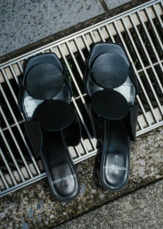 A pair of black Pierre Hardy leather slide sandals with circular strap embellishments placed on an outdoor metal drainage grate.