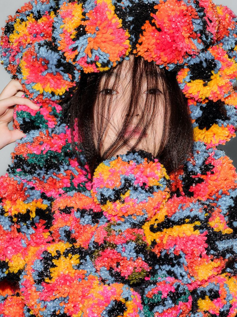 Asian woman wearing a multicolor pompom sweater and headpiece indoors against a grey wall; medium shot with fashion style.