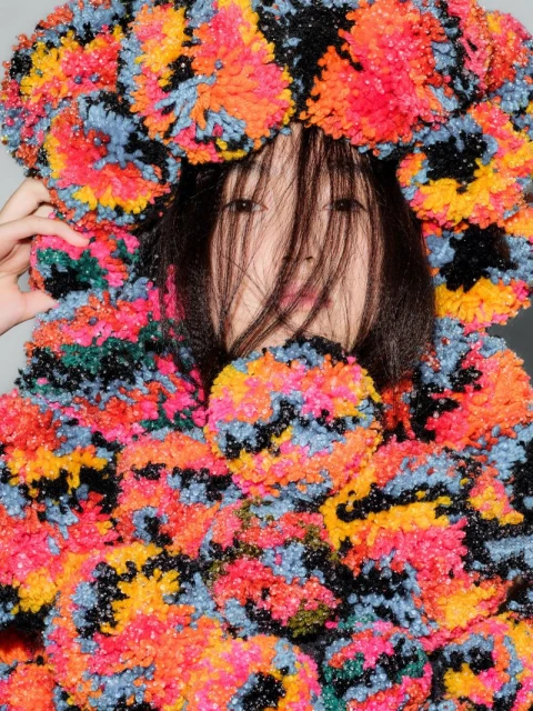 Asian woman wearing a multicolor pompom sweater and headpiece indoors against a grey wall; medium shot with fashion style.
