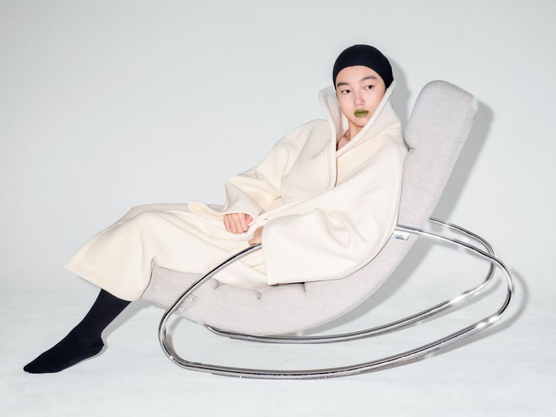 Woman with green lipstick in ivory robe poses in modern rocking chair against a white backdrop in a studio environment.