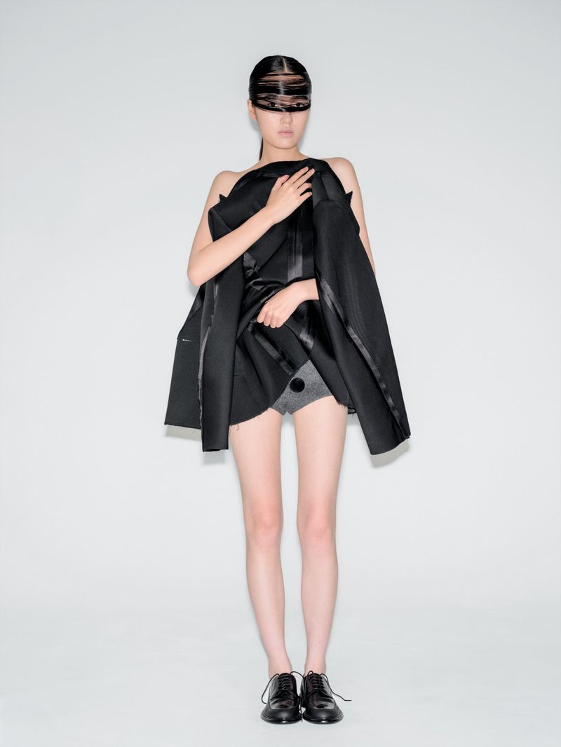 Asian woman with a futuristic black visor and avant-garde black blazer with minimal gray shorts and lace-up dress shoes in a studio.