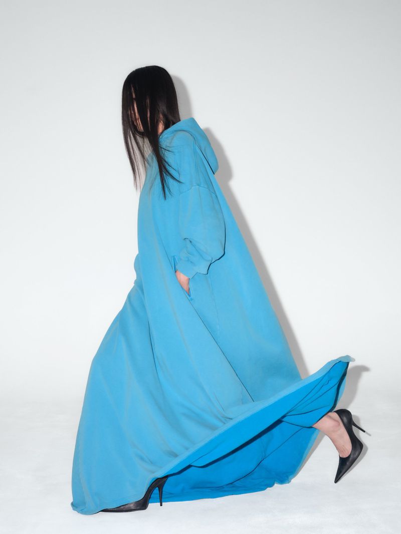 A woman in a long, blue, hooded dress and black high heels poses against a white backdrop, her face obscured by long dark hair.