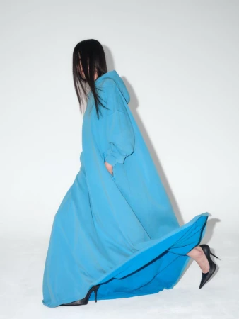 A woman in a long, blue, hooded dress and black high heels poses against a white backdrop, her face obscured by long dark hair.