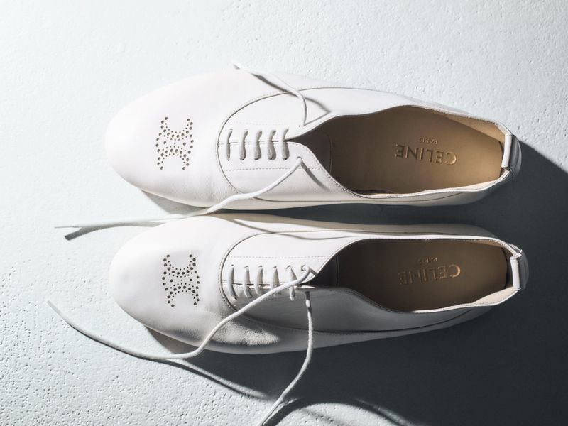 Overhead studio shot of two white leather Celine oxfords with laces on a light grey surface with shadow detail, showing the brand logo.