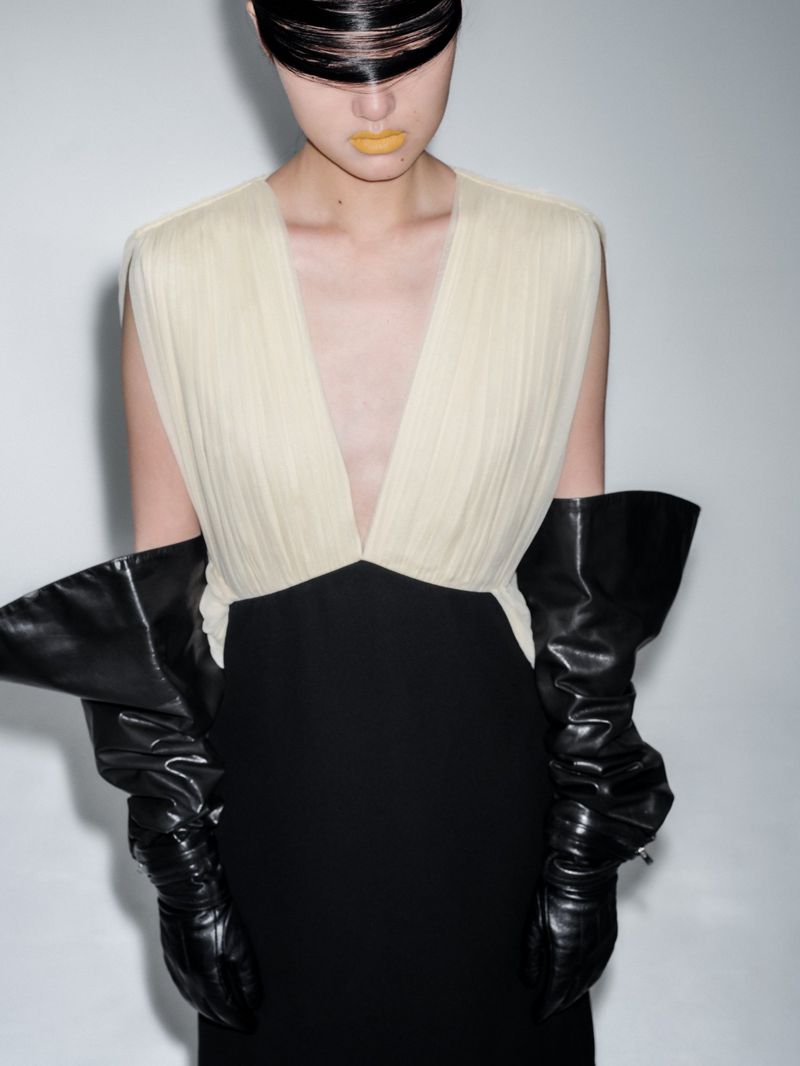 Woman in a modern black and white dress with high black shiny gloves poses against a bright white background in a studio fashion shoot.