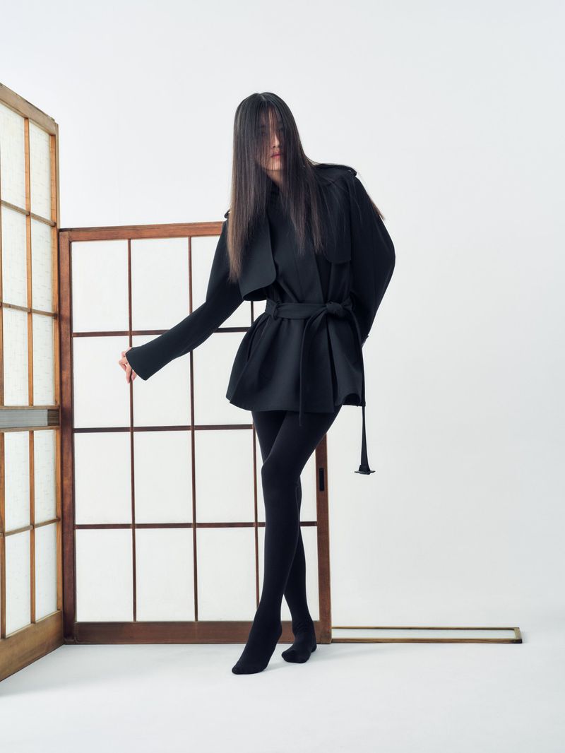 A solo adult female stands in a light studio wearing black coat, hosiery and heels next to a shoji screen and decorative beam.