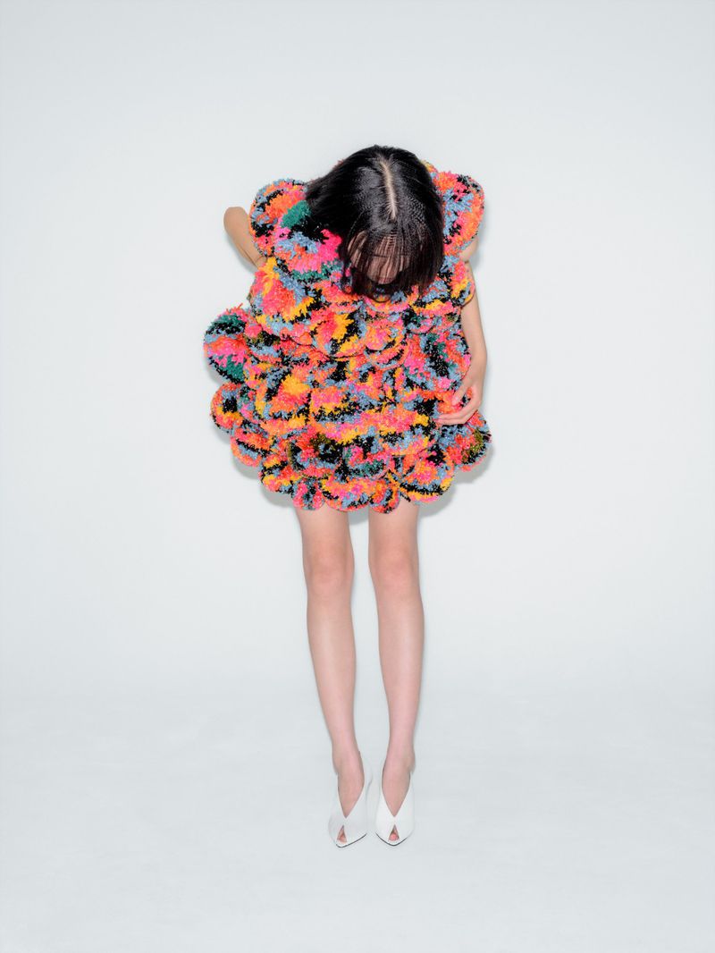 Woman in a colorful pompom dress and white high heels poses against a white wall, creating a vibrant fashion statement.