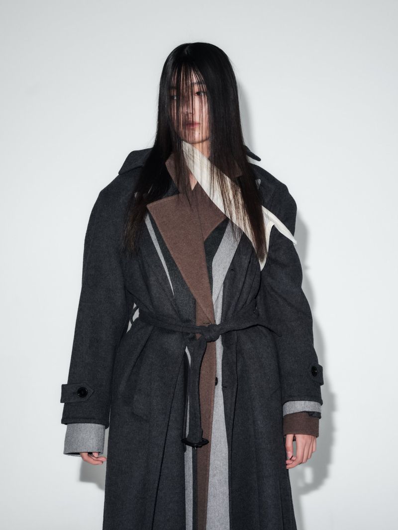 Model with long dark hair obscuring her face wearing a layered charcoal, brown, and grey wool overcoat against a white backdrop.