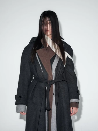 Model with long dark hair obscuring her face wearing a layered charcoal, brown, and grey wool overcoat against a white backdrop.