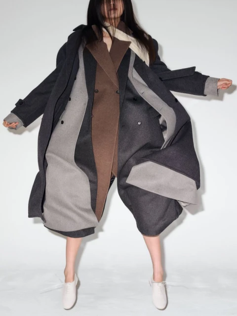 Woman in a brown and gray double-breasted coat posing in a studio with a white backdrop, looking down while floating in mid-air.