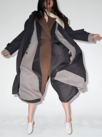 Woman in a brown and gray double-breasted coat posing in a studio with a white backdrop, looking down while floating in mid-air.