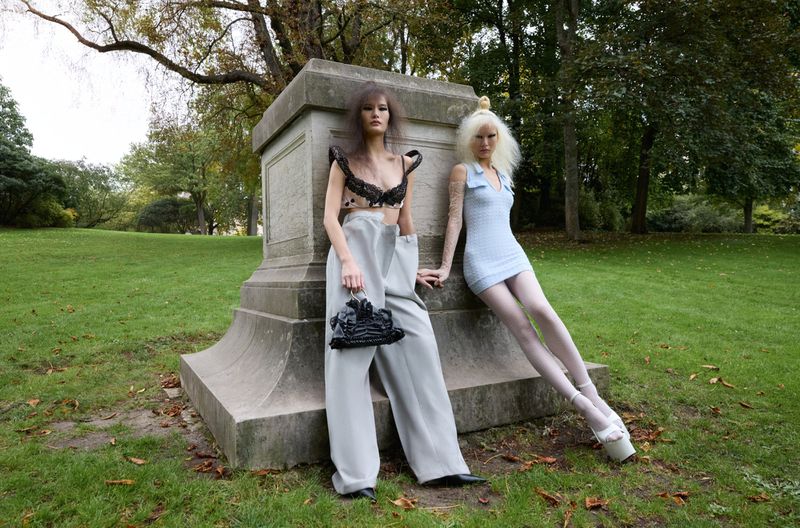 Two fashion models in avant-garde outfits posing against a large stone pedestal on a green park lawn.