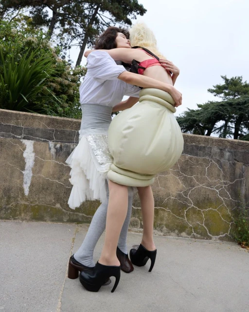 Two women embrace outside: one in a white shirt and skirt, the other in an unconventional garment with avant-garde platform shoes.