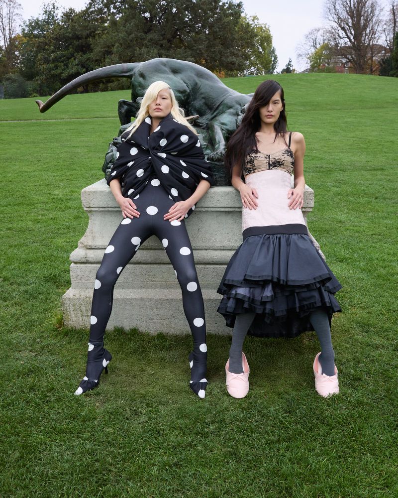 Two fashion models in polka dot leggings and tiered skirt pose near an outdoor sculpture on a green lawn with trees in the background.