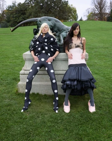 Two fashion models in polka dot leggings and tiered skirt pose near an outdoor sculpture on a green lawn with trees in the background.