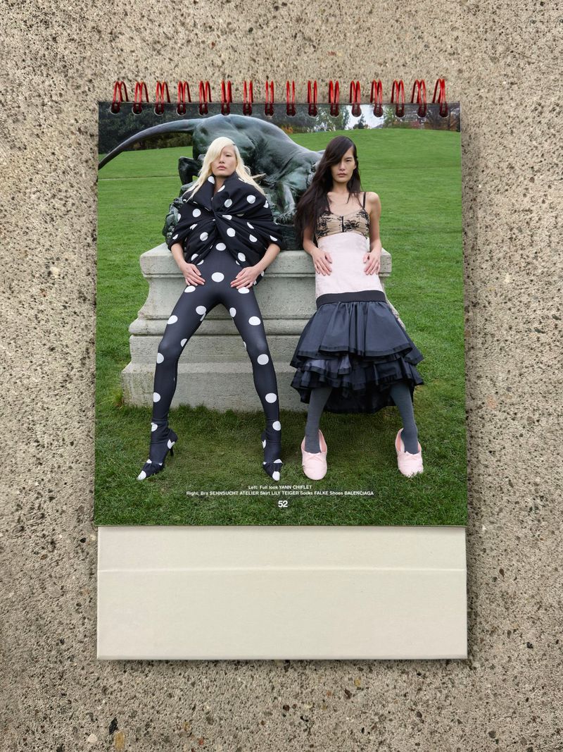 Two women pose in fashionable outfits in front of an outdoor sculpture on a calendar page, showcasing unique style and design.