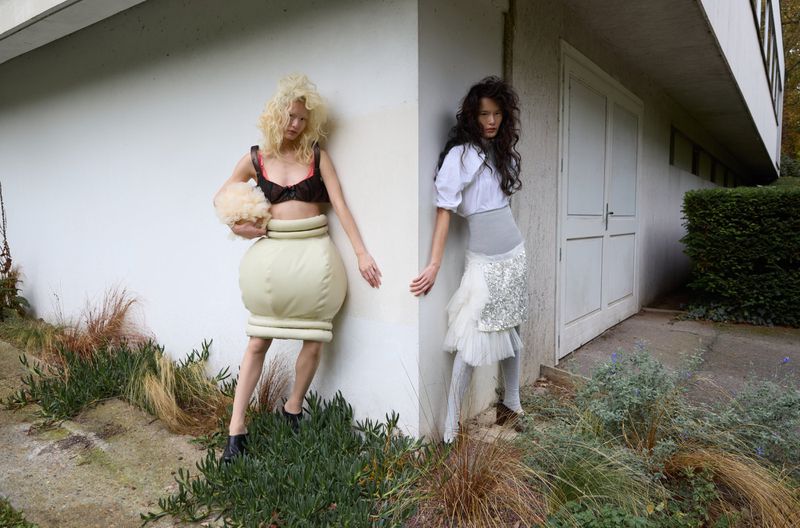Two women in unconventional fashion pose outdoors; one with blonde hair, one with dark hair, against a white building and green foliage.