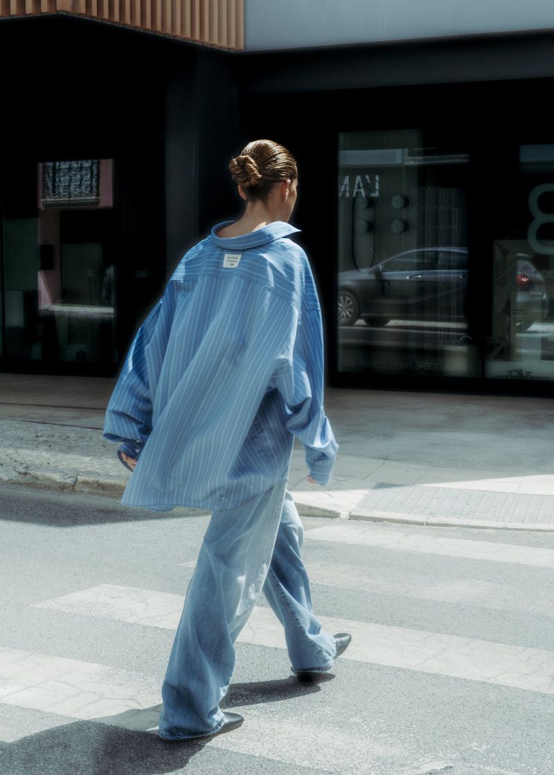 Back view of an adult female crossing a zebra-striped crosswalk in a city wearing a blue striped oversized shirt and baggy denim jeans.
