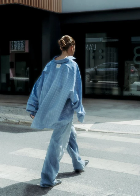 Back view of an adult female crossing a zebra-striped crosswalk in a city wearing a blue striped oversized shirt and baggy denim jeans.