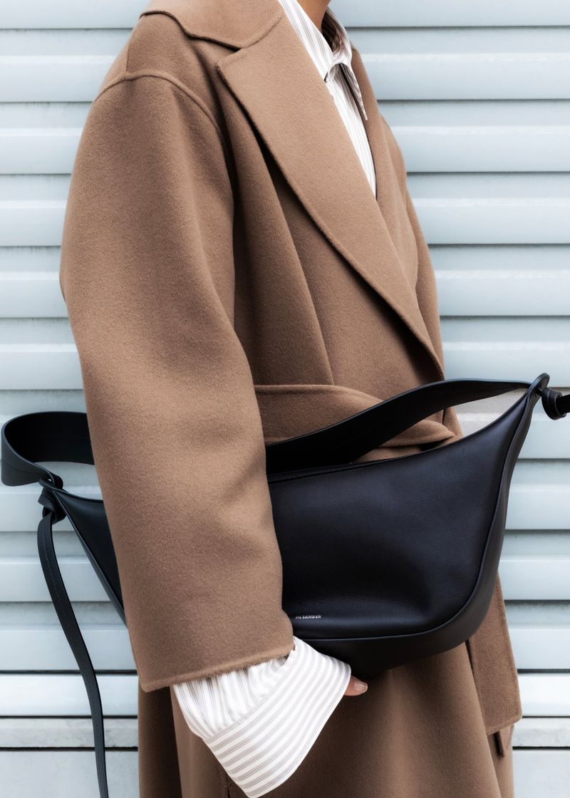 Fashion shot featuring a woman in a tan coat with a black shoulder bag, posed against a pale blue wall, showcasing style and elegance.