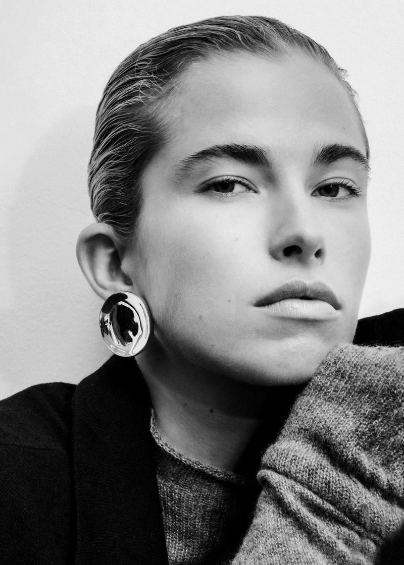 Black and white portrait of a woman with slicked-back hair wearing a large round silver earring and knit sweater.