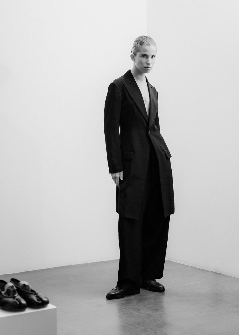 Black and white fashion shot of a slender adult posing in a dark suit, with black dress shoes displayed on a white block.