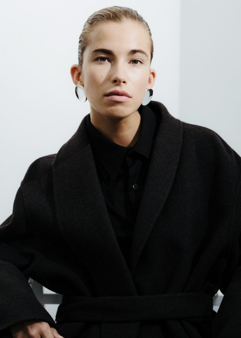 A poised woman with sleek, blonde hair poses indoors, wearing a dark belted wool coat and large earrings against a white backdrop.