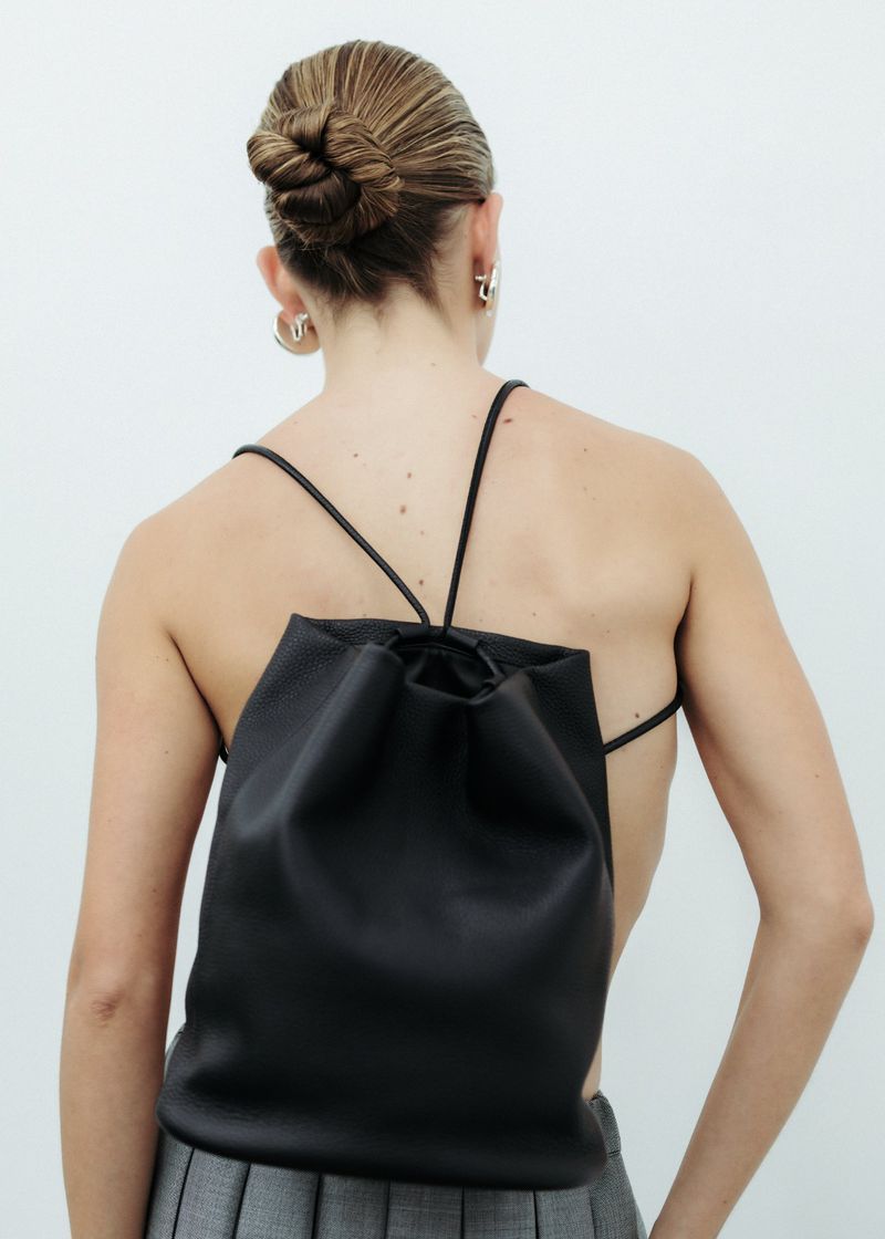 Back view of an adult female with a bun, wearing a black drawstring backpack and grey skirt against a white wall indoors.