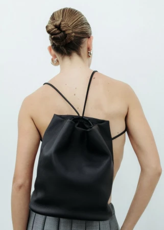 Back view of an adult female with a bun, wearing a black drawstring backpack and grey skirt against a white wall indoors.