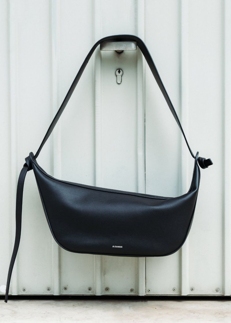 Studio shot of a black leather shoulder bag with a long strap hanging on a white corrugated metal background with a keyhole.