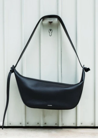 Studio shot of a black leather shoulder bag with a long strap hanging on a white corrugated metal background with a keyhole.